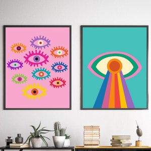May include: Two framed art prints. One features multiple colorful eye illustrations on a pink background. The other print displays a large eye with rainbow rays on a teal background. Both are in black frames.