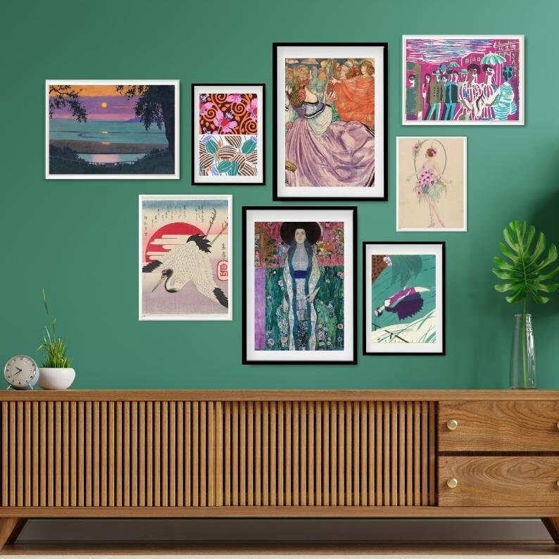 May include: A wall gallery featuring a variety of framed artwork, including a landscape scene, a floral design, a portrait of a woman, a Japanese-style crane illustration, and a group of people in a colourful scene. The artwork is displayed on a green wall above a wooden cabinet.