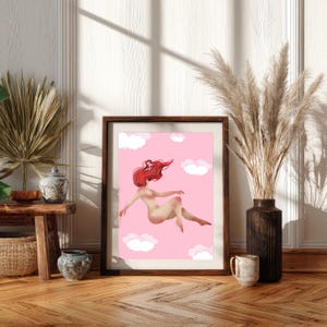 May include: Framed art print featuring a nude figure with flowing red hair floating among white clouds on a pink background. The artwork is displayed in a dark wooden frame, adding a touch of elegance to the whimsical scene. The print is set against a neutral wall and wooden floor.