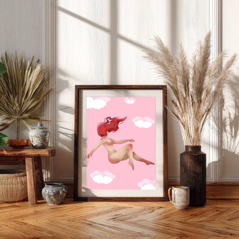 May include: Framed art print featuring a nude figure with flowing red hair floating among white clouds on a pink background. The artwork is displayed in a dark wooden frame, adding a touch of elegance to the whimsical scene. The print is set against a neutral wall and wooden floor.