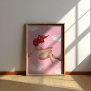 May include: A framed art print featuring a nude figure with flowing red hair floating in a pink sky with white clouds. The artwork is displayed in a light wood frame, leaning against a white wall.