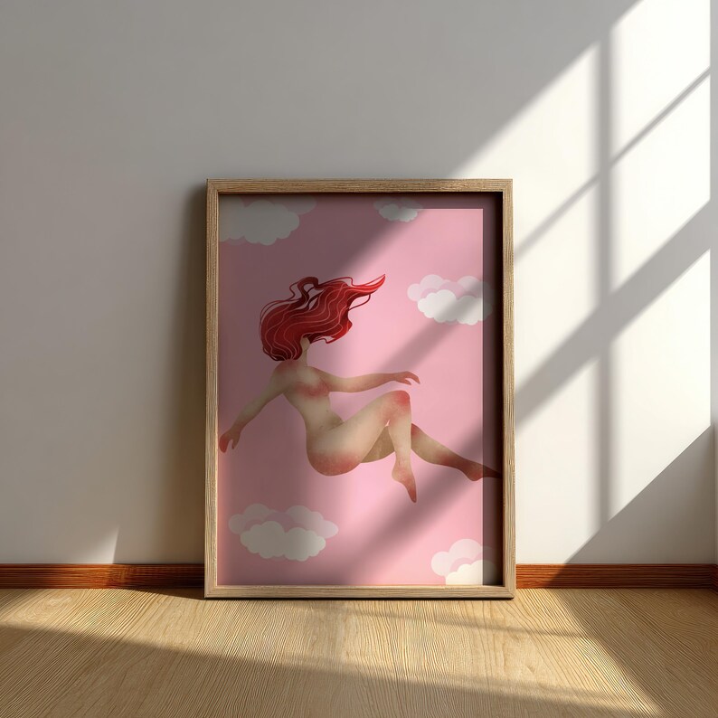 May include: A framed art print featuring a nude figure with flowing red hair floating in a pink sky with white clouds. The artwork is displayed in a light wood frame, leaning against a white wall.