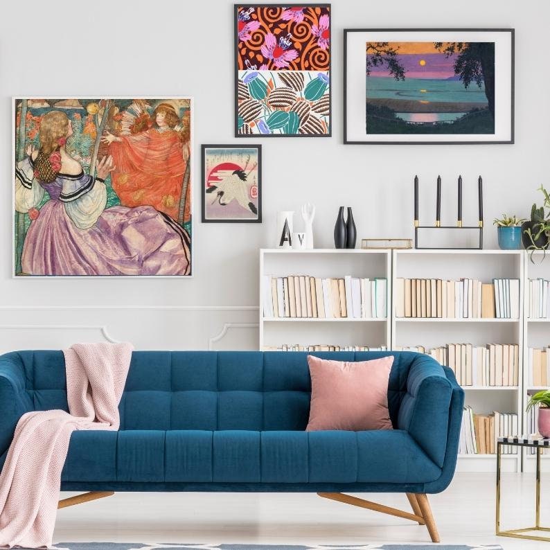 May include: A blue velvet sofa with a pink throw blanket and a pink cushion. The sofa is in front of a white wall with artwork and a bookshelf filled with books.