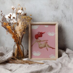 May include: A framed art print featuring a nude figure with flowing red hair floating among white clouds against a pink background. The artwork is in a light wooden frame. A vase of dried flowers and cotton stems sits to the left.