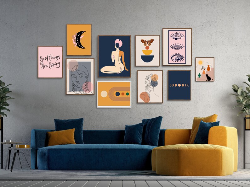 May include: A living room with a blue velvet sofa and a round ottoman. The wall behind the sofa is decorated with a gallery wall of framed prints featuring abstract designs, line art, and text. The prints are in various colors, including pink, blue, yellow, and orange. The text on one of the prints reads "Good things are coming."