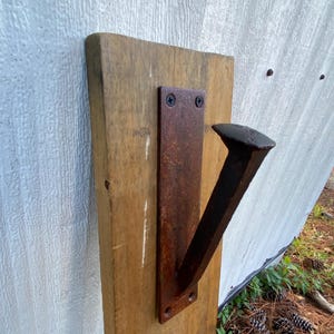 May include: A wall-mounted hook featuring a dark, rusted metal railroad spike attached to a rectangular metal plate, which is secured to a vertical, light-brown wooden board with visible wood grain and natural texture.