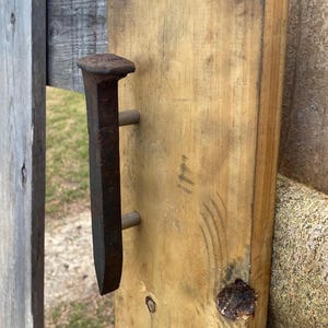 May include: A rustic wooden gate with a large, weathered, dark brown metal handle. The handle is attached to a light brown wooden plank with two metal pins. The gate is made of weathered wood and is outdoors.