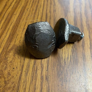 May include: Two dark gray, metal cabinet knobs with a textured, hammered appearance. The knobs have a rounded, faceted head and a square base. They are likely used for furniture or cabinet hardware.