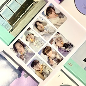 May include: A set of seven photo cards featuring seven different members of a K-pop group. Each card has a white border and features a close-up photo of a member wearing a winter hat and holding a lollipop. The cards are arranged in a grid pattern on a white background.