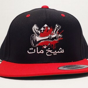 Puede incluir: Gorra de béisbol negra y roja con un diseño bordado blanco de una pieza de ajedrez con una salpicadura de sangre roja y el texto árabe "شبيح مات" en blanco.