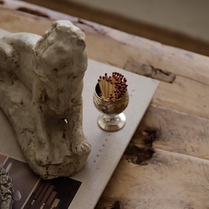 Pode incluir: Uma composi&ccedil;&atilde;o de natureza morta com um pequeno c&aacute;lice dourado cheio de f&oacute;sforos. Uma escultura texturizada de cor clara est&aacute; colocada ao lado do c&aacute;lice sobre um livro. A cena &eacute; montada sobre uma superf&iacute;cie de madeira r&uacute;stica.