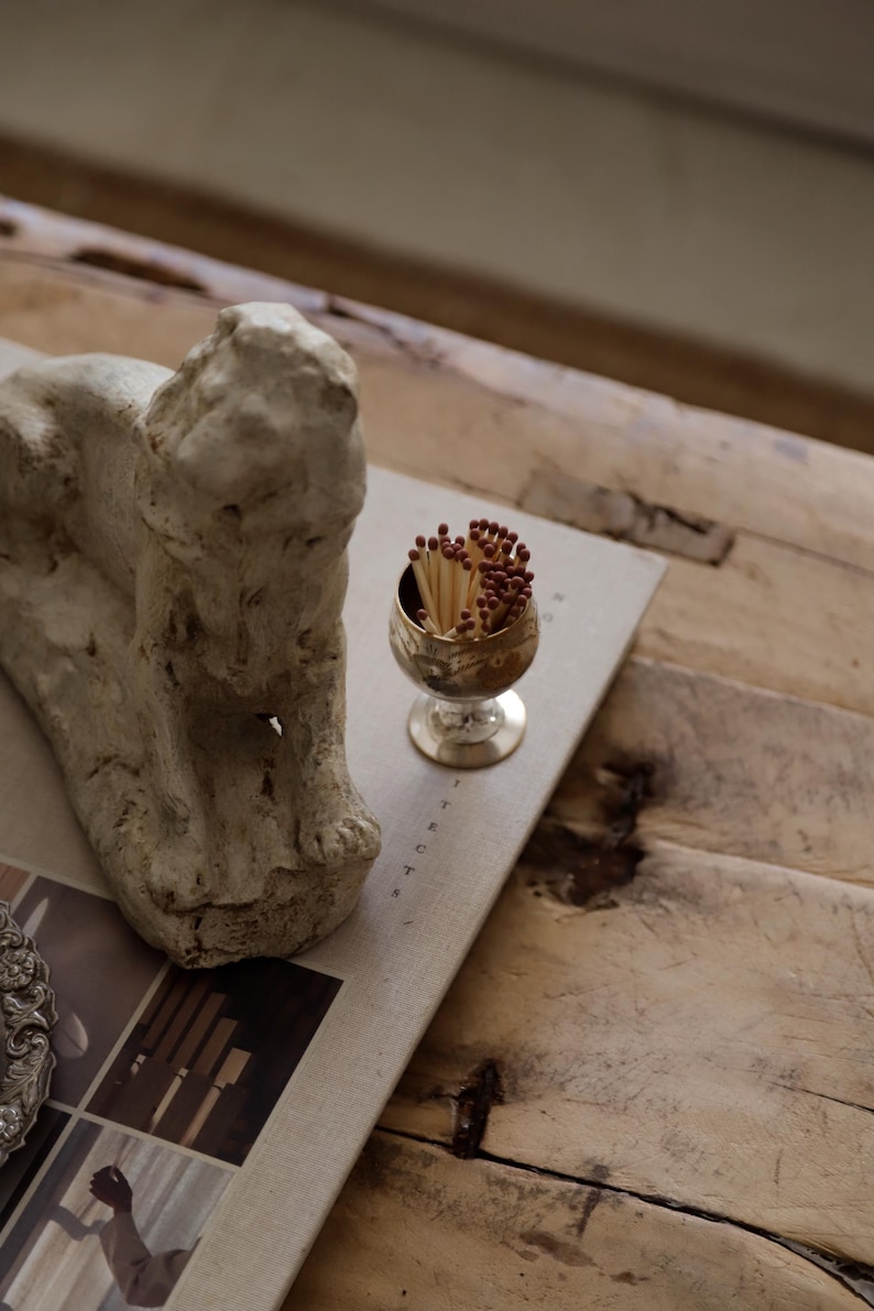 Pode incluir: Uma composi&ccedil;&atilde;o de natureza morta com um pequeno c&aacute;lice dourado cheio de f&oacute;sforos. Uma escultura texturizada de cor clara est&aacute; colocada ao lado do c&aacute;lice sobre um livro. A cena &eacute; montada sobre uma superf&iacute;cie de madeira r&uacute;stica.