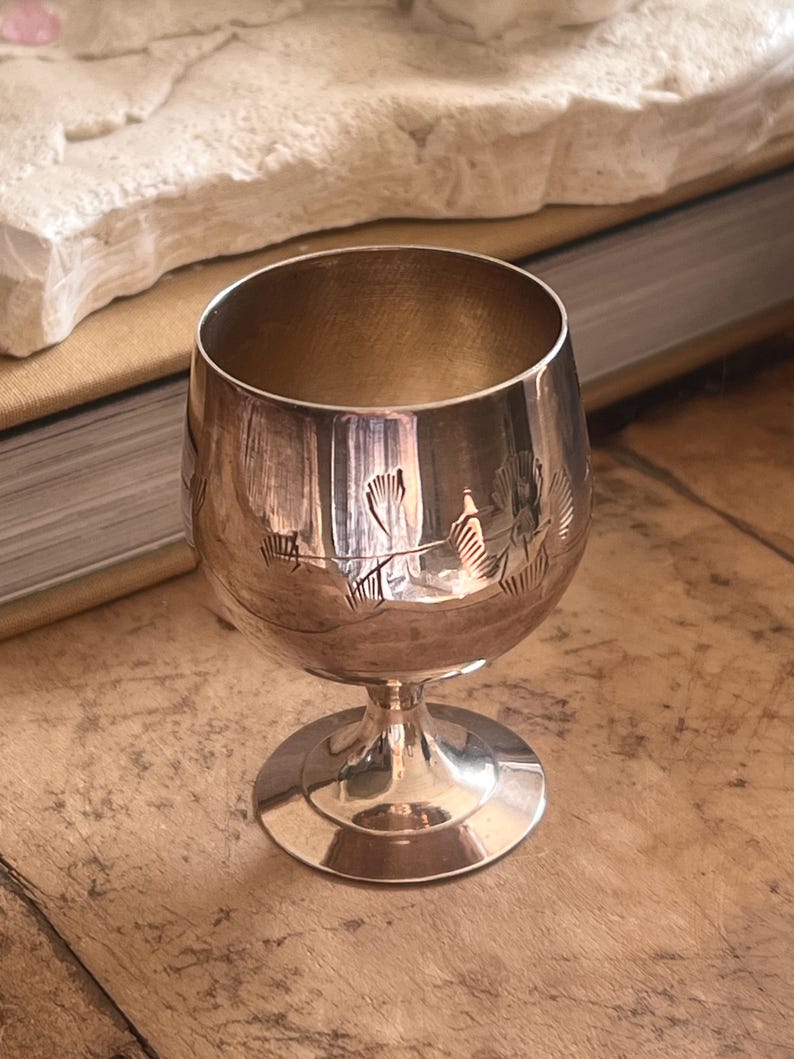 Pode incluir: Um pequeno c&aacute;lice prateado com base redonda e haste. A ta&ccedil;a de vidro tem um desenho gravado no meio. O c&aacute;lice est&aacute; sobre uma superf&iacute;cie de madeira.