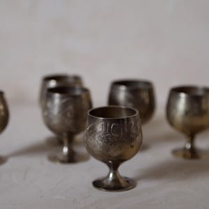 Pode incluir: Uma cole&ccedil;&atilde;o de pequenos c&aacute;lices de prata antigos com desenhos gravados detalhados. Os c&aacute;lices t&ecirc;m uma forma de ta&ccedil;a arredondada, suportada por uma haste curta e uma base circular. A prata tem uma apar&ecirc;ncia ligeiramente manchada, adicionando ao seu charme vintage.
