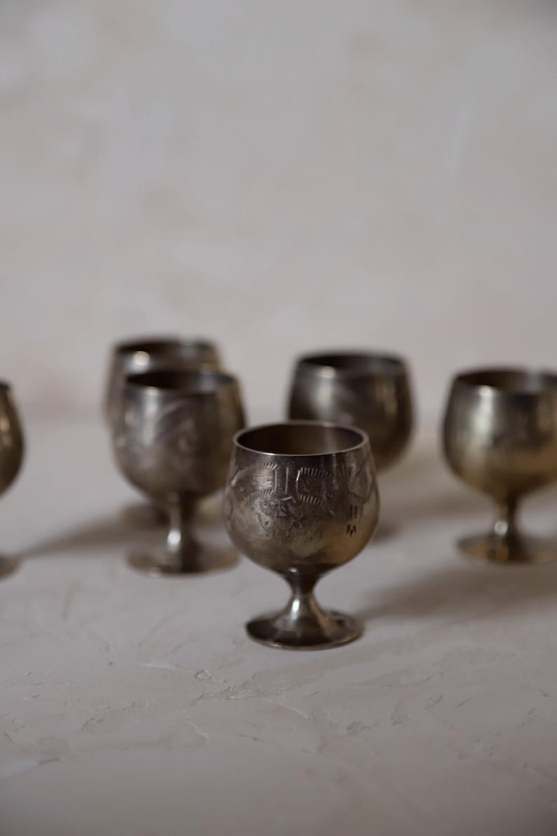 Pode incluir: Uma cole&ccedil;&atilde;o de pequenos c&aacute;lices de prata antigos com desenhos gravados detalhados. Os c&aacute;lices t&ecirc;m uma forma de ta&ccedil;a arredondada, suportada por uma haste curta e uma base circular. A prata tem uma apar&ecirc;ncia ligeiramente manchada, adicionando ao seu charme vintage.