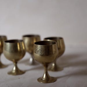Pode incluir: Uma cole&ccedil;&atilde;o de c&aacute;lices pequenos, cor de lat&atilde;o. Cada c&aacute;lice tem uma ta&ccedil;a arredondada, uma haste fina e uma base circular. Um c&aacute;lice tem um desenho gravado perto da borda. Os c&aacute;lices est&atilde;o dispostos sobre uma superf&iacute;cie branca.