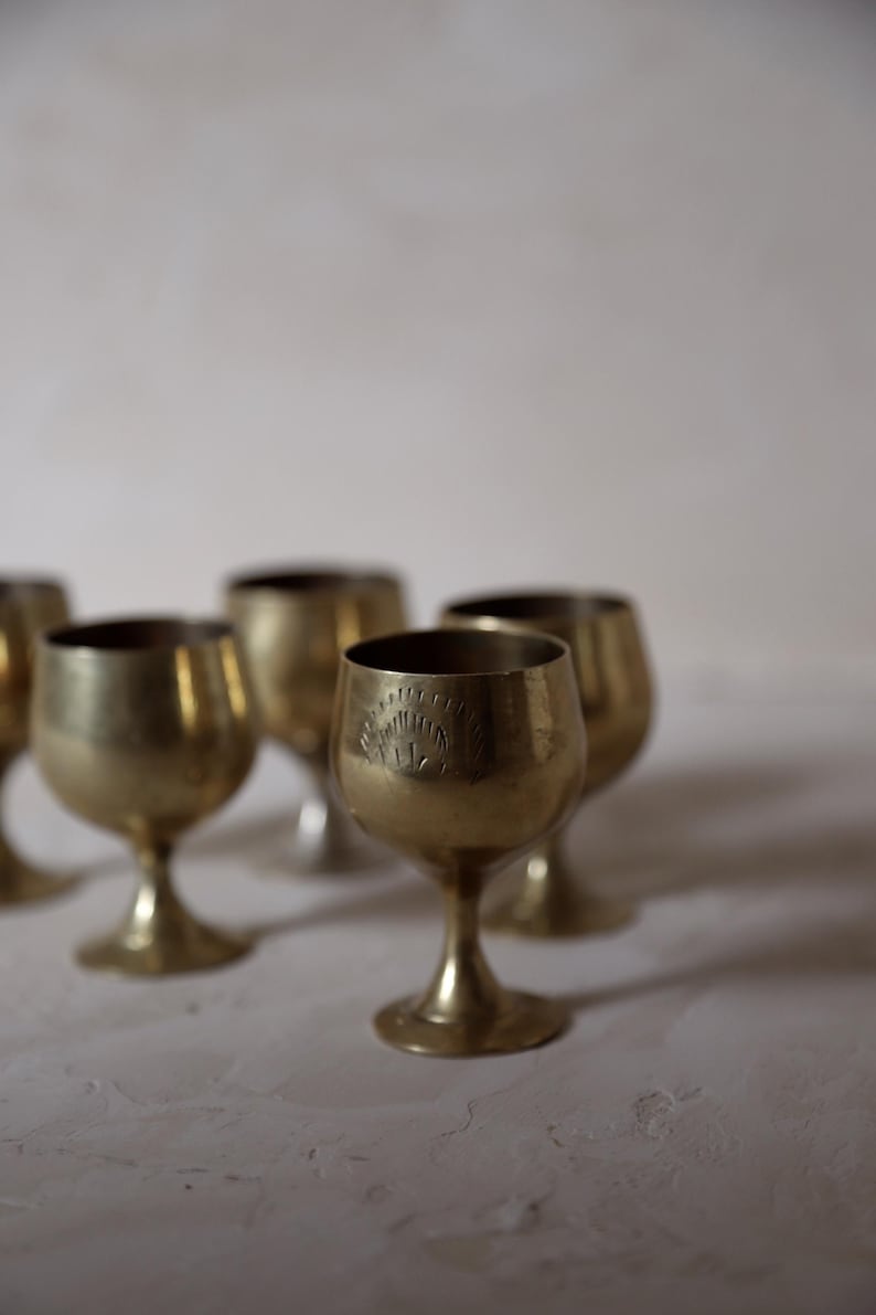 Pode incluir: Uma cole&ccedil;&atilde;o de c&aacute;lices pequenos, cor de lat&atilde;o. Cada c&aacute;lice tem uma ta&ccedil;a arredondada, uma haste fina e uma base circular. Um c&aacute;lice tem um desenho gravado perto da borda. Os c&aacute;lices est&atilde;o dispostos sobre uma superf&iacute;cie branca.