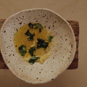 May include: A speckled, off-white ceramic bowl holds a pool of yellow oil, garnished with fresh green herbs. The bowl sits on a small, weathered wooden surface, creating a rustic aesthetic. The food preparation is simple and elegant.