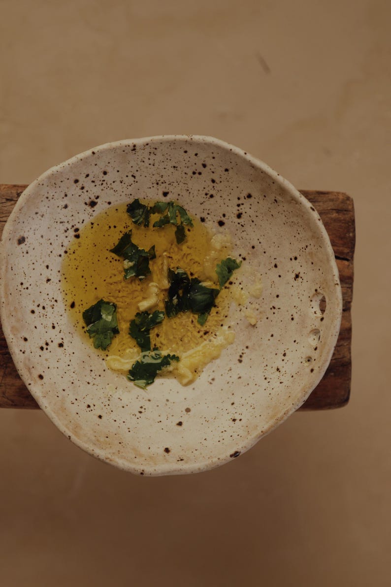 May include: A speckled, off-white ceramic bowl holds a pool of yellow oil, garnished with fresh green herbs. The bowl sits on a small, weathered wooden surface, creating a rustic aesthetic. The food preparation is simple and elegant.