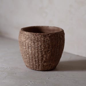 Puede incluir: Una maceta de cerámica marrón con textura tejida. La maceta es redonda y tiene una abertura ancha.