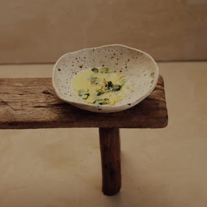 May include: A speckled, off-white ceramic bowl filled with a yellow liquid and green herbs, resting on a rustic wooden stool. The stool has a single, round leg. The background is a neutral, light brown color.