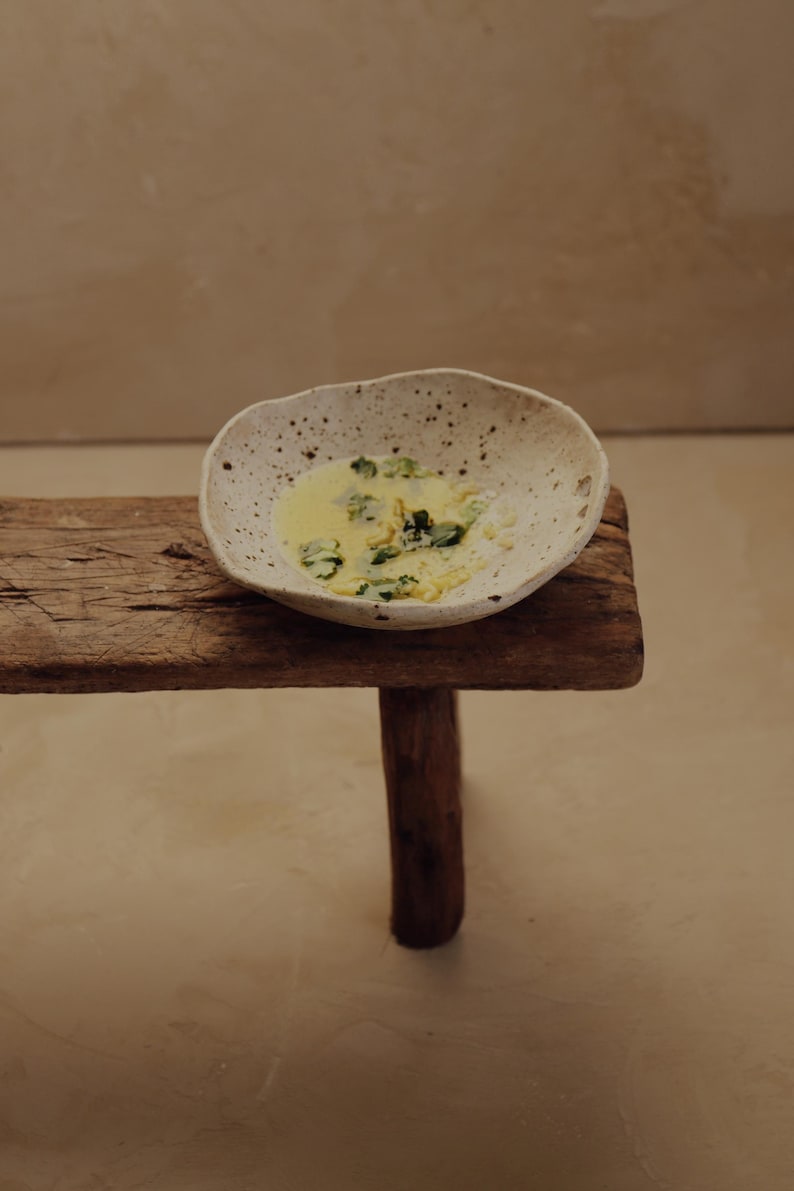 May include: A speckled, off-white ceramic bowl filled with a yellow liquid and green herbs, resting on a rustic wooden stool. The stool has a single, round leg. The background is a neutral, light brown color.