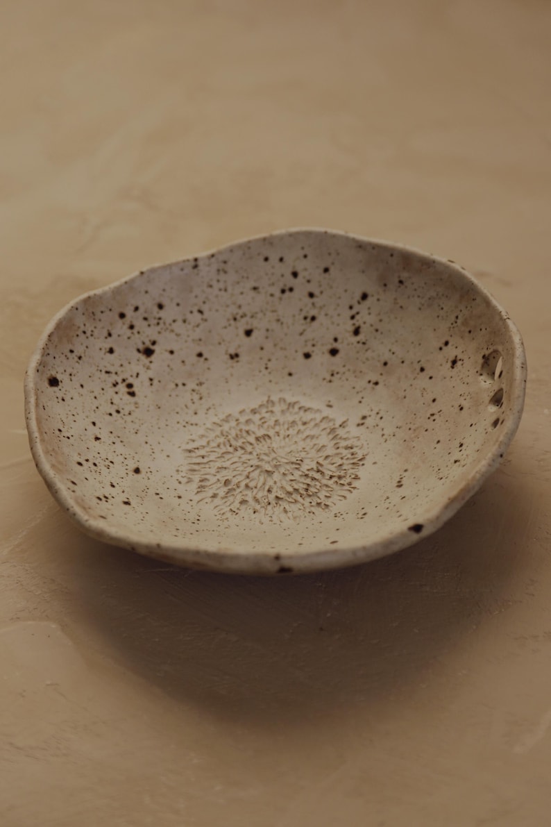May include: A speckled, off-white ceramic bowl with an organic shape. The bowl features a textured, floral-like design in the center and small holes near the rim. The bowl is likely used for serving food or as a decorative piece.