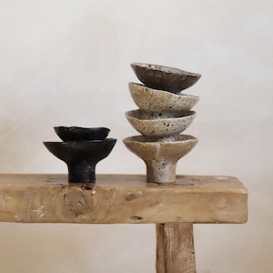 Puede incluir: Colección de pequeños cuencos de cerámica hechos a mano en tonos negro, beige y marrón moteado. Los cuencos están apilados y colocados sobre una superficie de madera rústica, mostrando sus formas y texturas únicas. La paleta de colores neutros crea una estética minimalista.