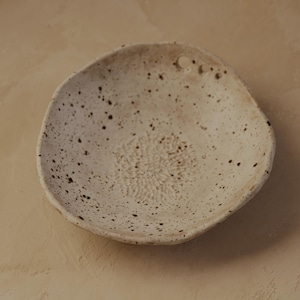 May include: A handmade, off-white ceramic bowl with a speckled pattern. The bowl has an organic shape with a textured interior and small, dark brown speckles throughout. The centre features a detailed, raised floral design.