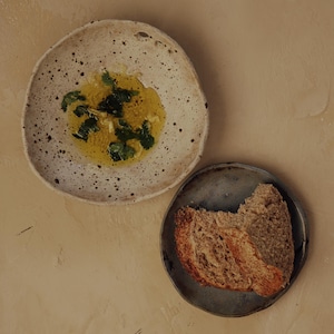 May include: Overhead shot of two ceramic plates on a beige surface. One plate holds olive oil with herbs, the other holds slices of bread. The plates are different sizes and colors.