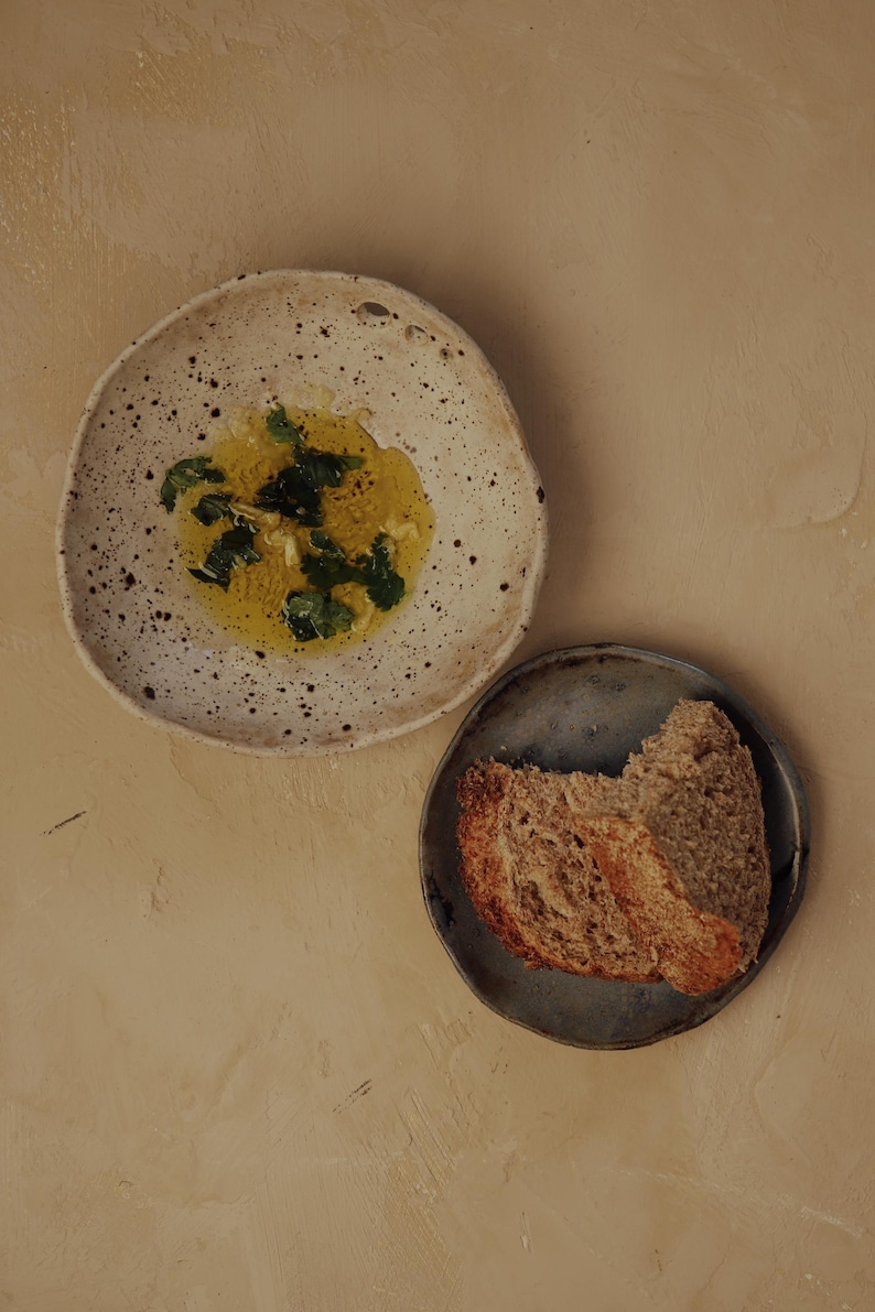May include: Overhead shot of two ceramic plates on a beige surface. One plate holds olive oil with herbs, the other holds slices of bread. The plates are different sizes and colors.