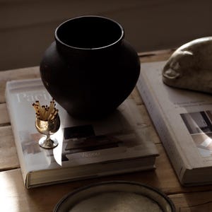Pode incluir: Uma natureza morta com um vaso preto fosco, um pequeno c&aacute;lice de prata cheio de f&oacute;sforos e dois livros. A palavra "Paci" &eacute; vis&iacute;vel em um dos livros. Uma bandeja de prata est&aacute; em primeiro plano. Os objetos est&atilde;o dispostos sobre uma superf&iacute;cie de madeira.
