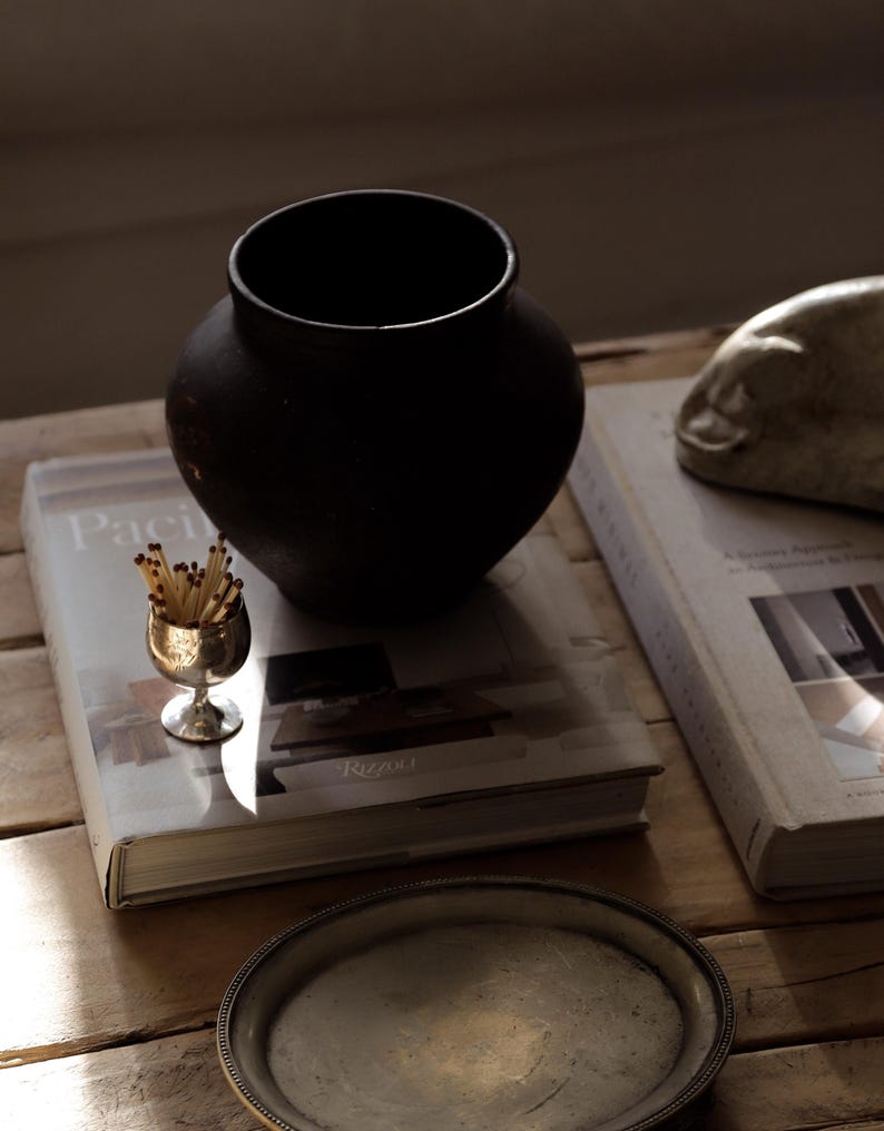 Pode incluir: Uma natureza morta com um vaso preto fosco, um pequeno c&aacute;lice de prata cheio de f&oacute;sforos e dois livros. A palavra "Paci" &eacute; vis&iacute;vel em um dos livros. Uma bandeja de prata est&aacute; em primeiro plano. Os objetos est&atilde;o dispostos sobre uma superf&iacute;cie de madeira.