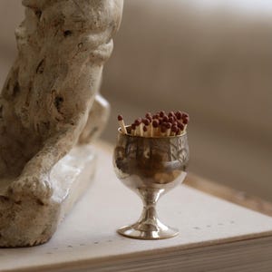 Pode incluir: Um pequeno c&aacute;lice prateado cheio de f&oacute;sforos de madeira. O c&aacute;lice est&aacute; sobre um livro grosso ao lado de uma escultura desgastada. Os f&oacute;sforos t&ecirc;m pontas castanhas. O c&aacute;lice tem um desenho gravado.