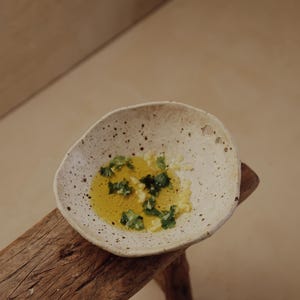 May include: A small, speckled ceramic bowl filled with olive oil, chopped herbs, and garlic. The bowl sits on a rustic wooden stool, with a neutral background.