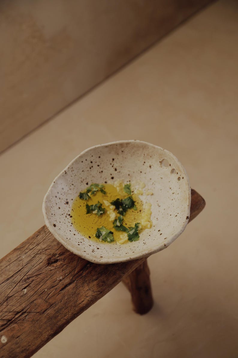 May include: A small, speckled ceramic bowl filled with olive oil, chopped herbs, and garlic. The bowl sits on a rustic wooden stool, with a neutral background.
