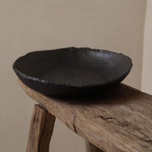 May include: A handcrafted, matte black ceramic bowl with a rough, textured rim. The bowl is placed on a rustic, weathered wooden stool. The bowl's dark color contrasts with the light wood, highlighting its unique, artisanal quality. Ideal for serving or as a decorative piece.