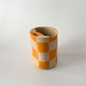 Könnte beinhalten: Ein Keramik-Zahnbürstenhalter mit einem lebendigen orangefarbenen und cremefarbenen Schachbrettmuster. Der zylindrische Halter hat einen kleinen, gebogenen Griff an der Seite und eine glatte, glänzende Oberfläche. Der Innenraum ist einfarbig cremefarben.