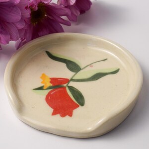 Peut inclure: Repose-cuillère en céramique crème avec un motif floral rouge et jaune. Le repose-cuillère a une forme incurvée avec une petite indentation pour reposer les ustensiles. Des feuilles vertes et une fleur rouge sont peintes sur la surface.