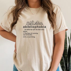 Pu&ograve; includere: Maglietta beige con la parola "abibliophobia" e la sua pronuncia in nero. Sotto, una definizione recita: "La paura di rimanere senza libri. &Egrave; una cosa reale."
