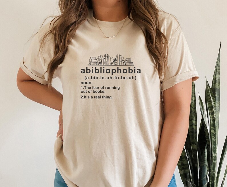 Pu&ograve; includere: Maglietta beige con la parola "abibliophobia" e la sua pronuncia in nero. Sotto, una definizione recita: "La paura di rimanere senza libri. &Egrave; una cosa reale."