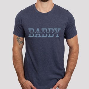 May include: A navy blue t-shirt with the word "DADDY" in a varsity style font. The names "JACK", "SOPHIA", "EMLY", and "JOHN" are printed below the word "DADDY" with a heart symbol between each name.