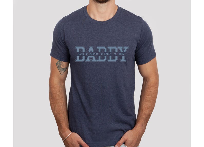 May include: A navy blue t-shirt with the word "DADDY" in a varsity style font. The names "JACK", "SOPHIA", "EMLY", and "JOHN" are printed below the word "DADDY" with a heart symbol between each name.