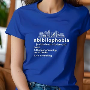 Pu&ograve; includere: T-shirt blu reale con la parola "abibliophobia" e la sua definizione stampata in bianco. La definizione include la pronuncia, la parte del discorso e una descrizione della paura di rimanere senza libri.