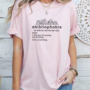 Pu&ograve; includere: T-shirt rosa chiaro con la parola "abibliophobia" stampata in nero, insieme alla sua definizione. La maglietta presenta un disegno di una pila di libri sopra la parola. La definizione afferma la paura di rimanere senza libri.