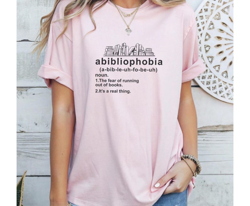 Pu&ograve; includere: T-shirt rosa chiaro con la parola "abibliophobia" stampata in nero, insieme alla sua definizione. La maglietta presenta un disegno di una pila di libri sopra la parola. La definizione afferma la paura di rimanere senza libri.