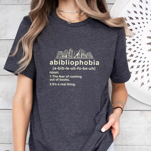 Pu&ograve; includere: T-shirt grigio scuro con la parola "abibliophobia" e la sua pronuncia tra parentesi. Sotto, la definizione recita: "1. La paura di rimanere senza libri. 2. &Egrave; una cosa reale." Una grafica di libri &egrave; sopra la parola.