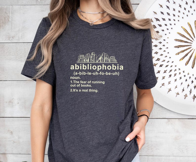 Pu&ograve; includere: T-shirt grigio scuro con la parola "abibliophobia" e la sua pronuncia tra parentesi. Sotto, la definizione recita: "1. La paura di rimanere senza libri. 2. &Egrave; una cosa reale." Una grafica di libri &egrave; sopra la parola.