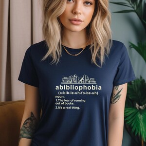 Pu&ograve; includere: T-shirt blu navy con la parola "abibliophobia" e la sua definizione stampate in bianco. Il design include una grafica di libri impilati. Il testo della maglietta definisce l'abibliofobia come la paura di rimanere senza libri.
