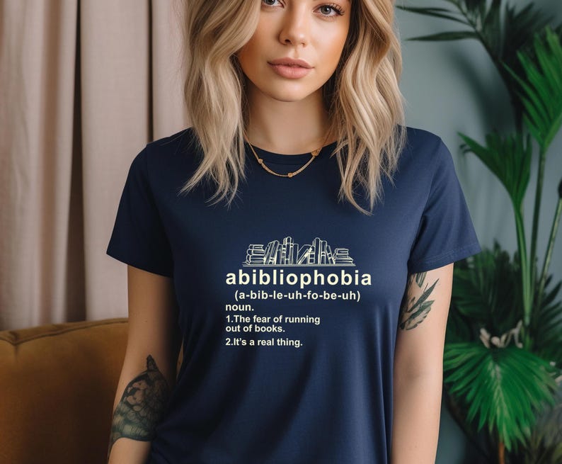 Pu&ograve; includere: T-shirt blu navy con la parola "abibliophobia" e la sua definizione stampate in bianco. Il design include una grafica di libri impilati. Il testo della maglietta definisce l'abibliofobia come la paura di rimanere senza libri.